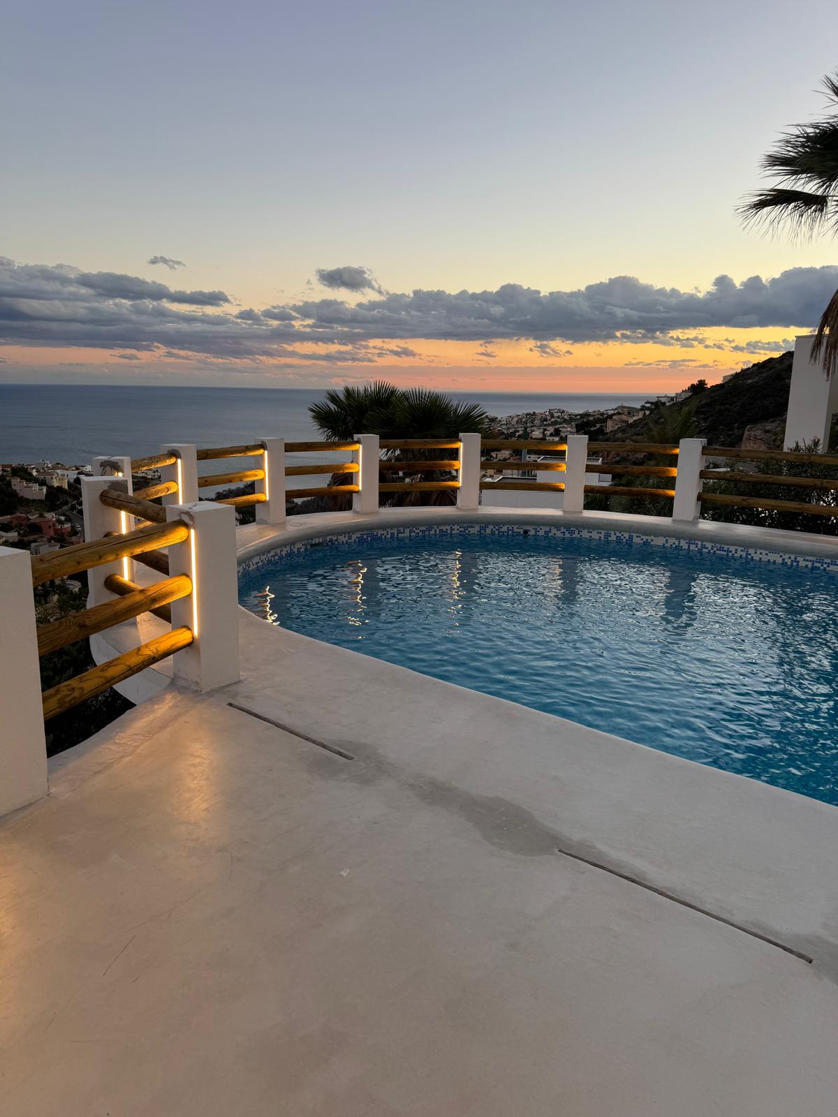 Piscina circular con gresite azul e iluminación LED integrada en la barandilla de madera, ofreciendo vistas panorámicas al mar durante el atardecer.