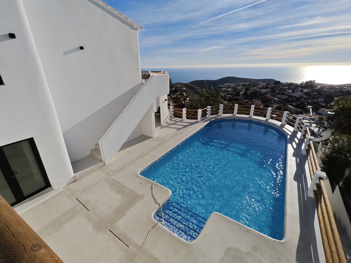 Piscina de mosaico azul y terraza de cemento blanco con barandillas de madera, ofreciendo vistas panorámicas al mar Mediterráneo y el entorno costero.