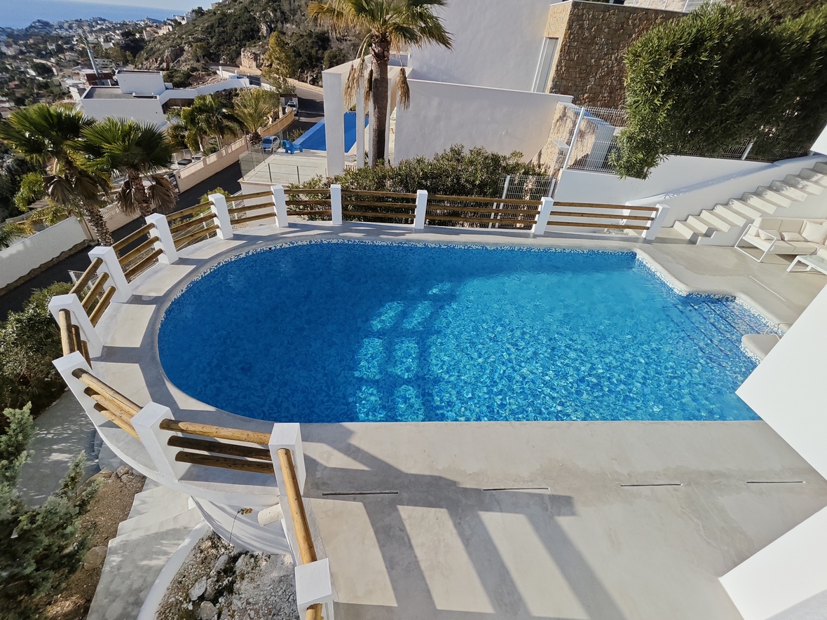 Piscina exterior curva con gresite azul, rodeada por una terraza de piedra y barandilla de madera con vistas panorámicas a la costa mediterránea.