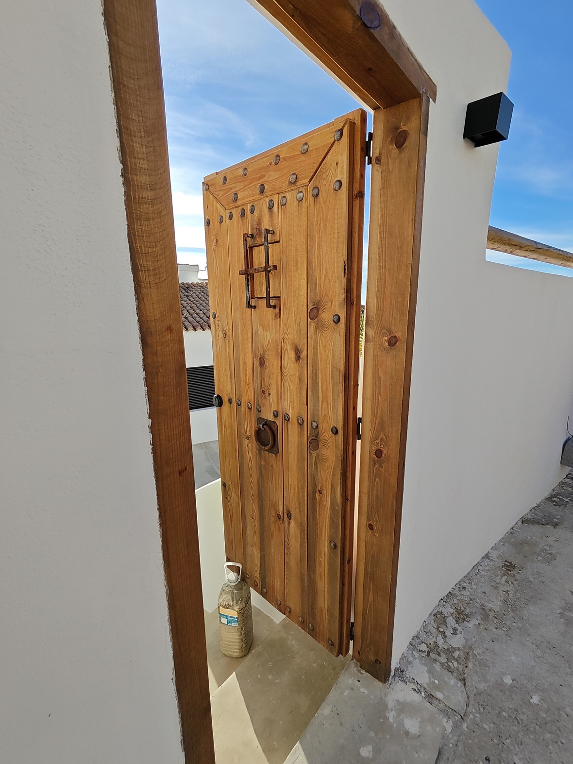 Puerta rústica de madera con clavos de hierro y mirilla enrejada, integrada en una estructura de paredes blancas y marco de madera maciza.