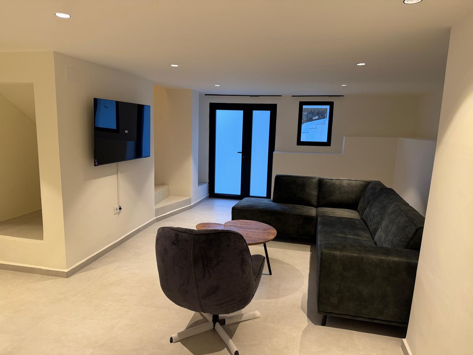 Sala de estar con sofá en L oscuro, sillón giratorio y TV de pared. Incluye iluminación empotrada, suelos cerámicos neutros y acceso exterior con carpintería negra.