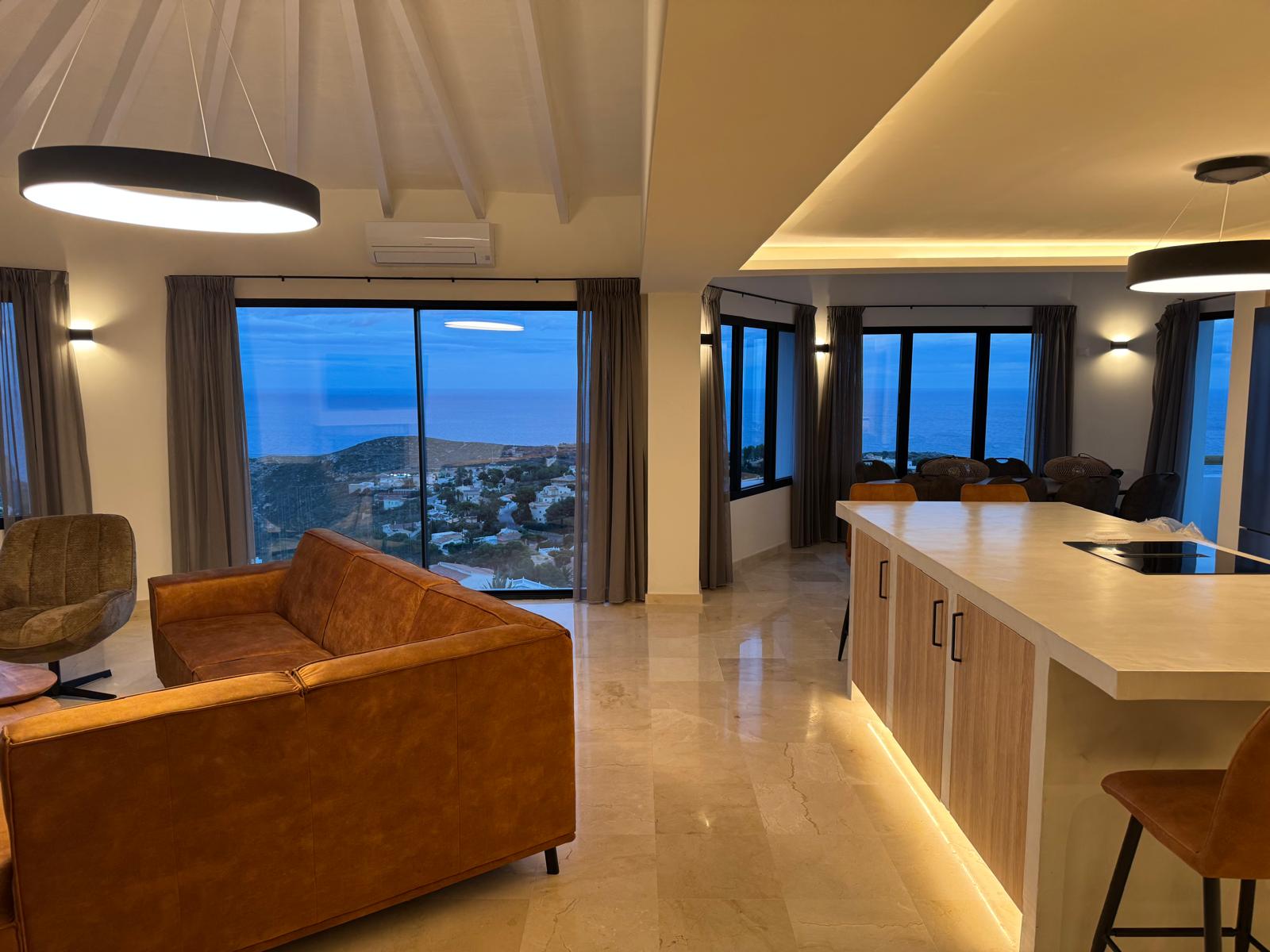Salón de planta abierta con sofá de cuero color coñac e isla de cocina iluminada, ofreciendo vistas panorámicas al mar a través de amplios ventanales.