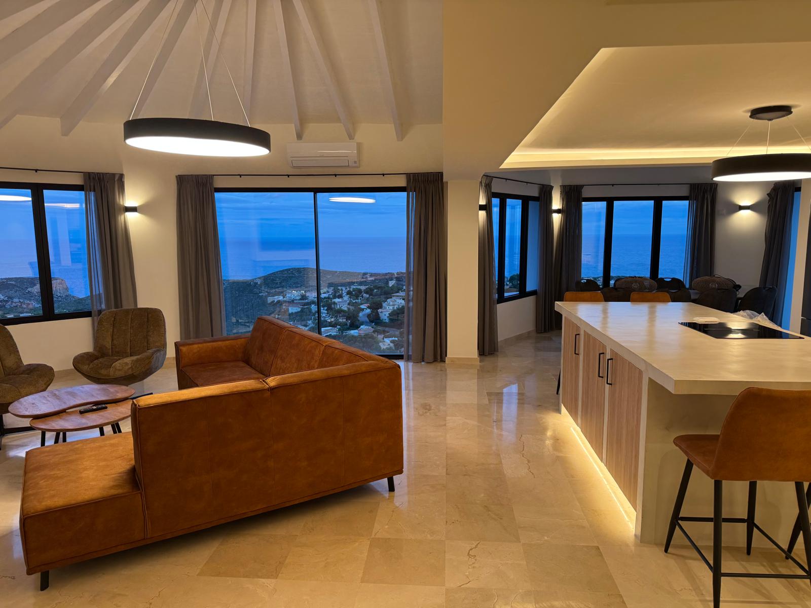Salón de planta abierta con sofá de cuero color coñac, isla de cocina con iluminación LED y amplios ventanales con vistas panorámicas al mar al anochecer.