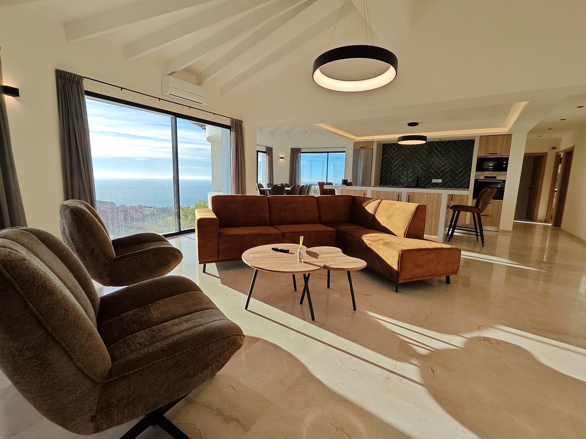 Salón de planta abierta con sofá en L color terracota, techos abovedados con vigas y ventanales con vistas al mar sobre suelos de mármol pulido.