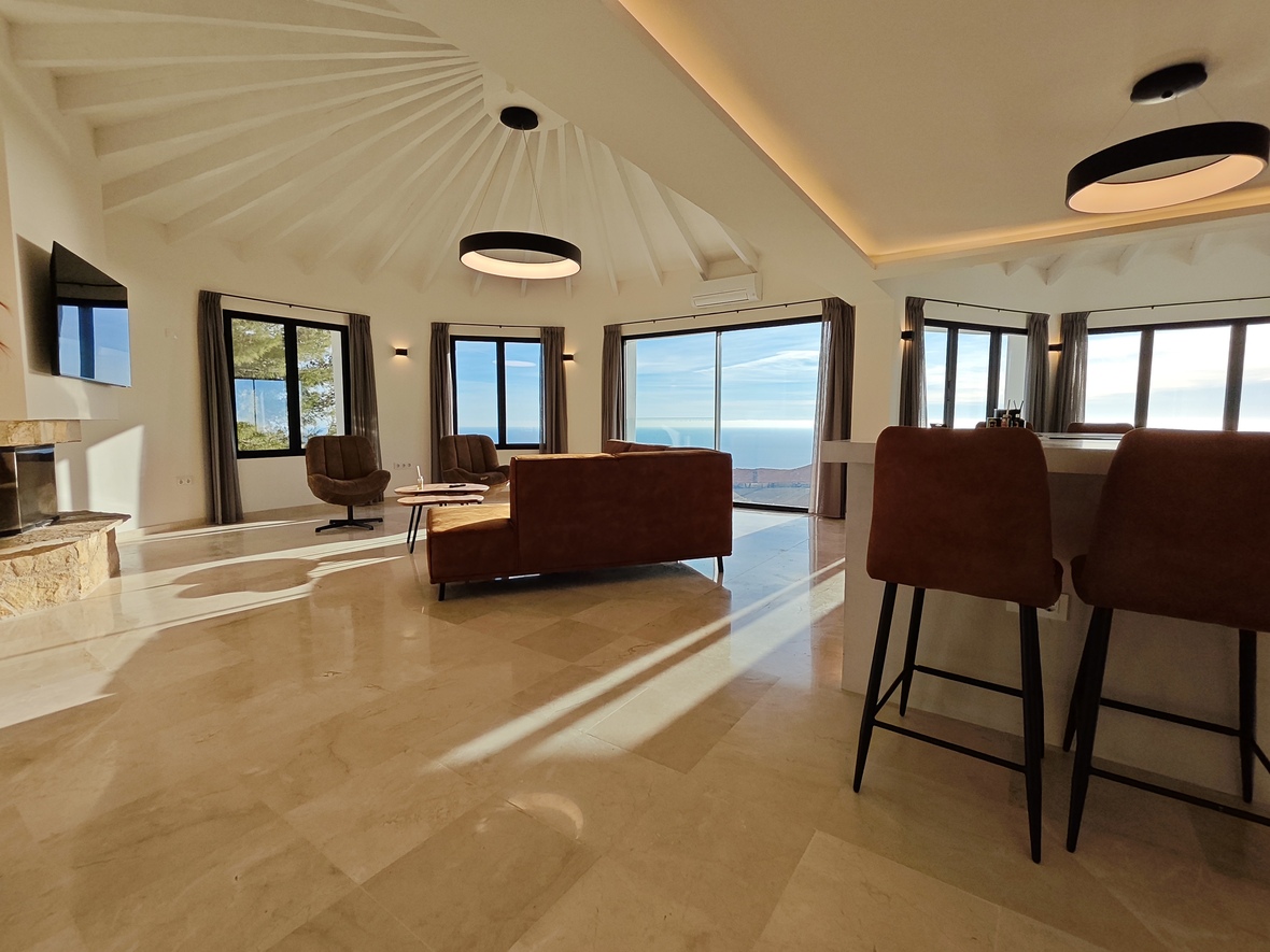 Salón diáfano con techo radial de vigas blancas, suelos de mármol pulido y vistas panorámicas al mar. Dispone de chimenea de piedra y luminarias circulares modernas.