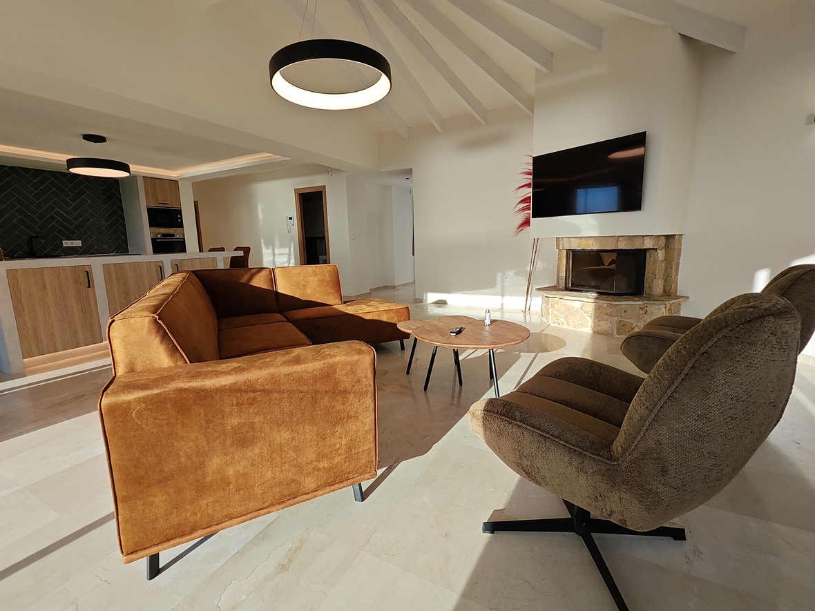 Salón moderno con sofá de terciopelo ocre, chimenea de piedra y techos abovedados con vigas. Al fondo destaca la cocina abierta con salpicadero de espiga verde y suelos de mármol.