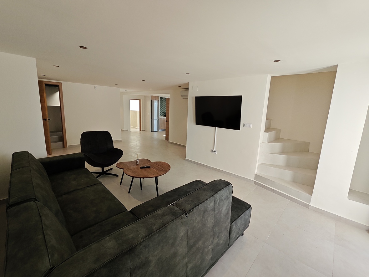 Salón moderno con sofá rinconera oscuro, mesa de centro de madera y TV de pared, junto a una escalera minimalista blanca e iluminación empotrada.