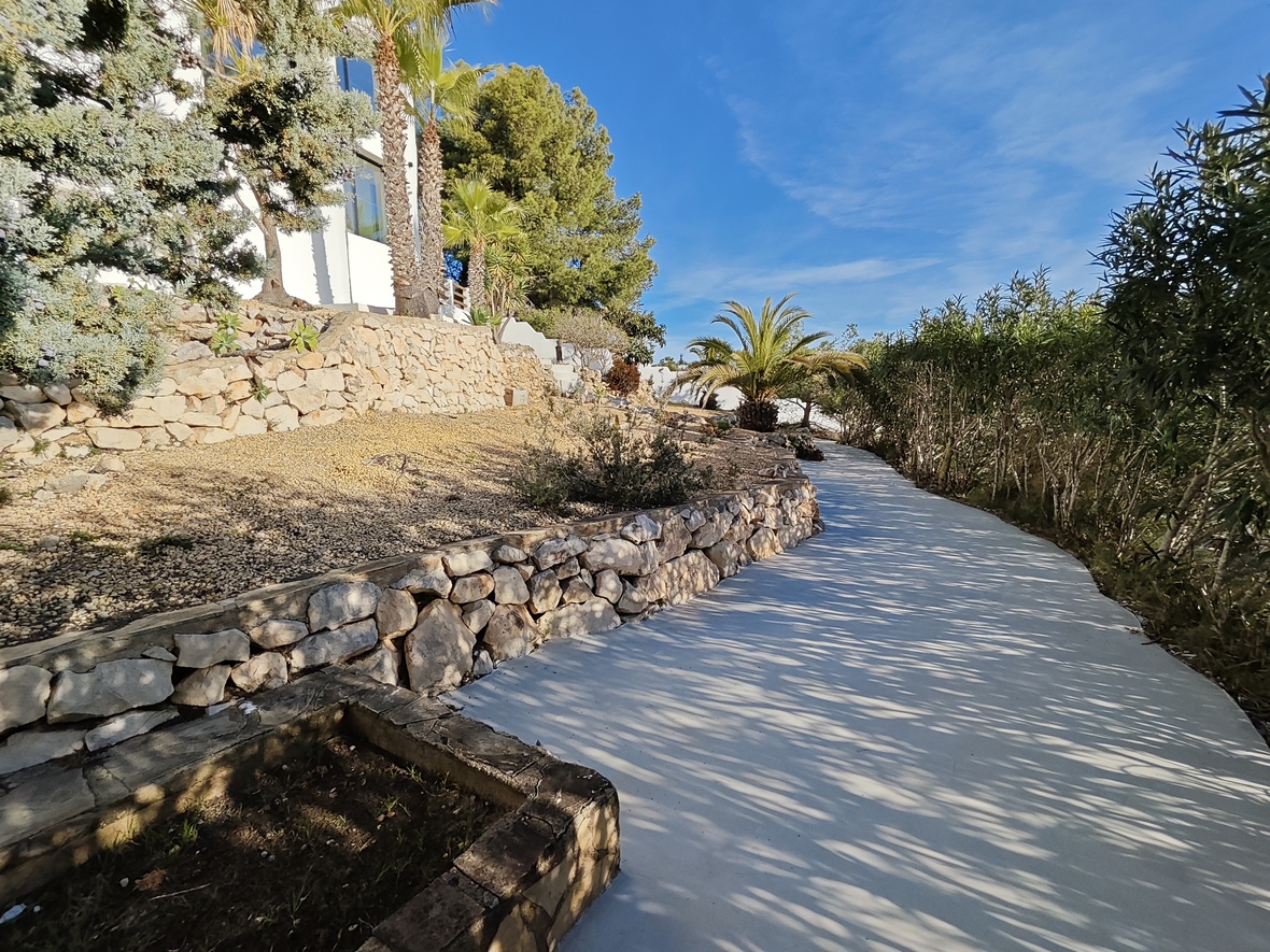 Sendero pavimentado que recorre un jardín mediterráneo con muros de contención de piedra seca, palmeras y pinos junto a la fachada blanca de la villa.