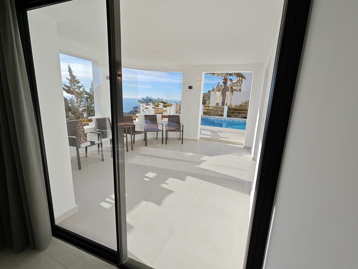 Terraza cubierta con suelos cerámicos claros y mobiliario exterior, ofreciendo vistas panorámicas al mar Mediterráneo y acceso visual a la piscina.