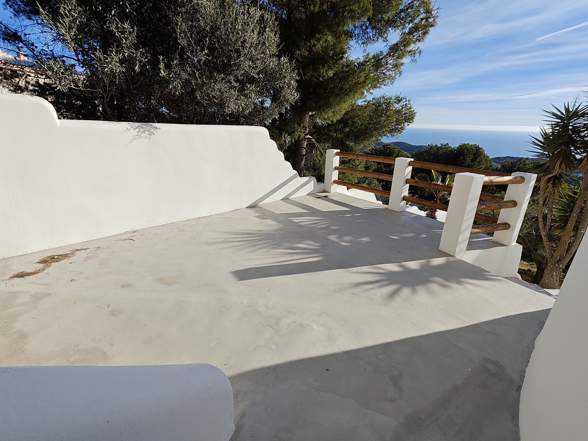 Terraza elevada con suelo de microcemento, muros de cal blanca y barandilla de madera con vistas panorámicas al mar Mediterráneo y al pinar circundante.
