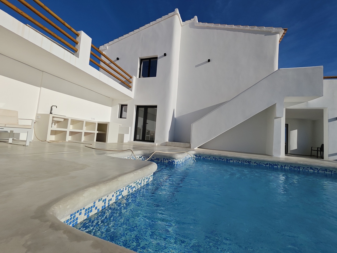Terraza exterior con piscina de gresite azul, cocina de verano integrada y fachada blanca minimalista con barandillas de madera y escalera.