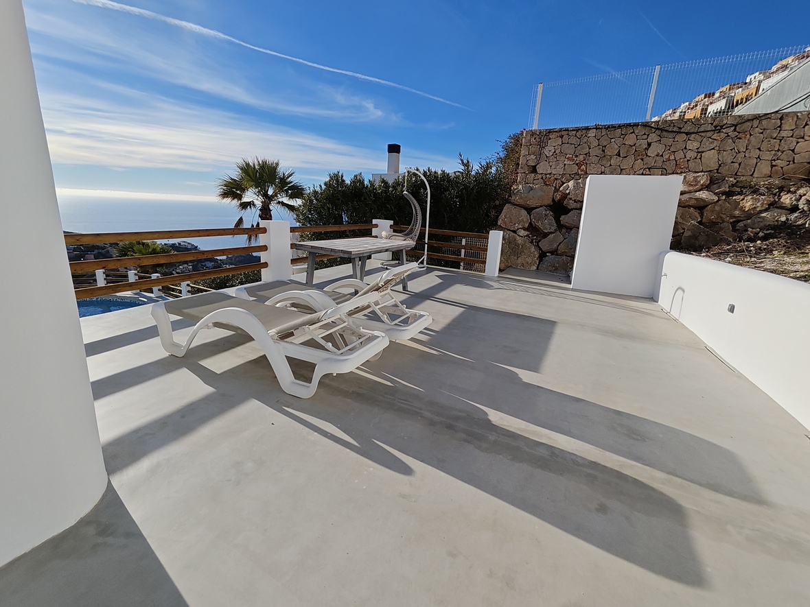 Terraza solárium con tumbonas blancas, mesa de madera y silla colgante. Ofrece vistas panorámicas al mar, muros de piedra natural y suelo de microcemento.