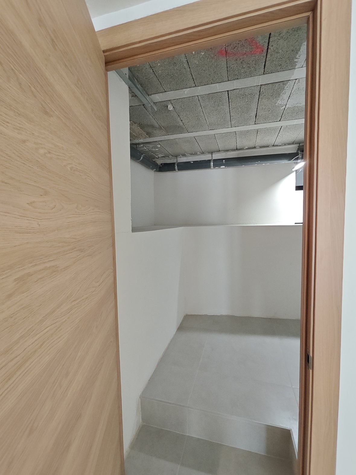 Vista a través de una puerta de madera clara hacia un cuarto trastero con suelo cerámico gris y techo técnico con tuberías de drenaje vistas.