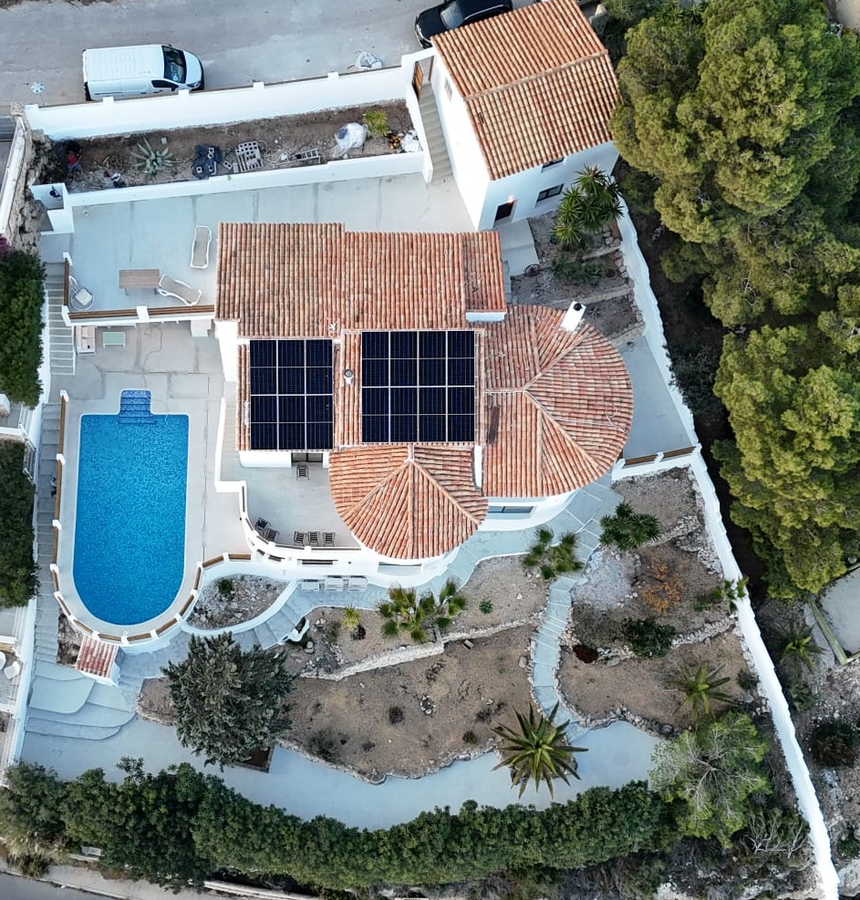 Vista aérea de una villa mediterránea con paneles solares en el tejado, piscina de forma curva y jardines aterrazados con vegetación autóctona.