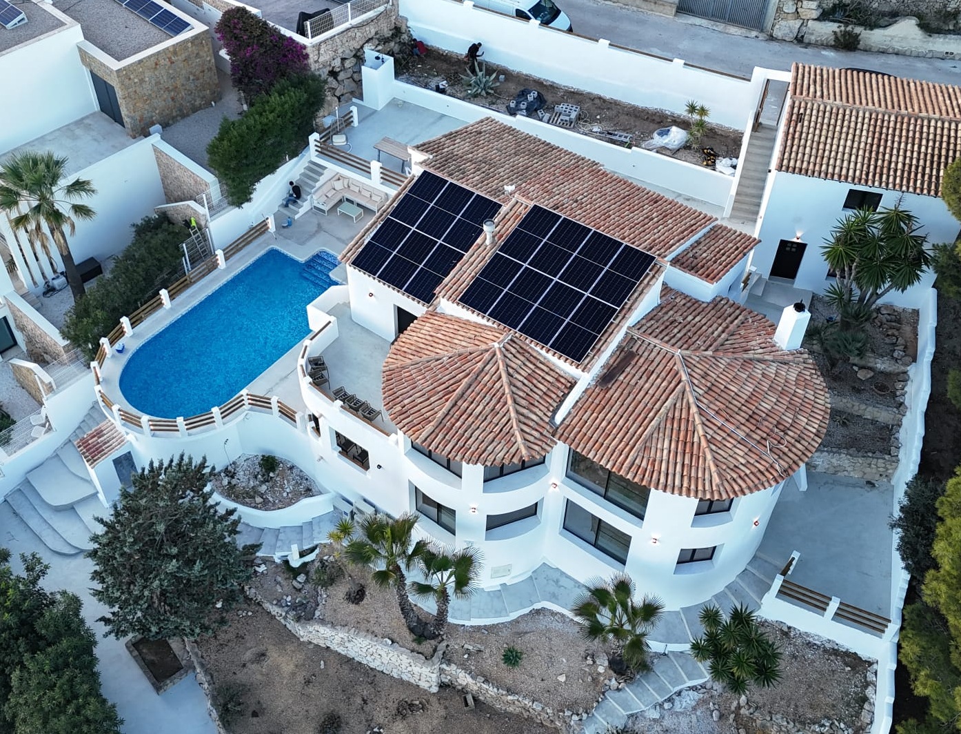 Vista aérea de una villa mediterránea con tejados de terracota, paneles solares, piscina ovalada y terrazas de piedra integradas en el terreno escalonado.