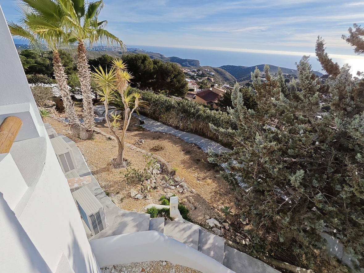 Vista elevada de un jardín con palmeras y senderos de grava, que ofrece una panorámica del valle costero y el mar Mediterráneo en el horizonte.