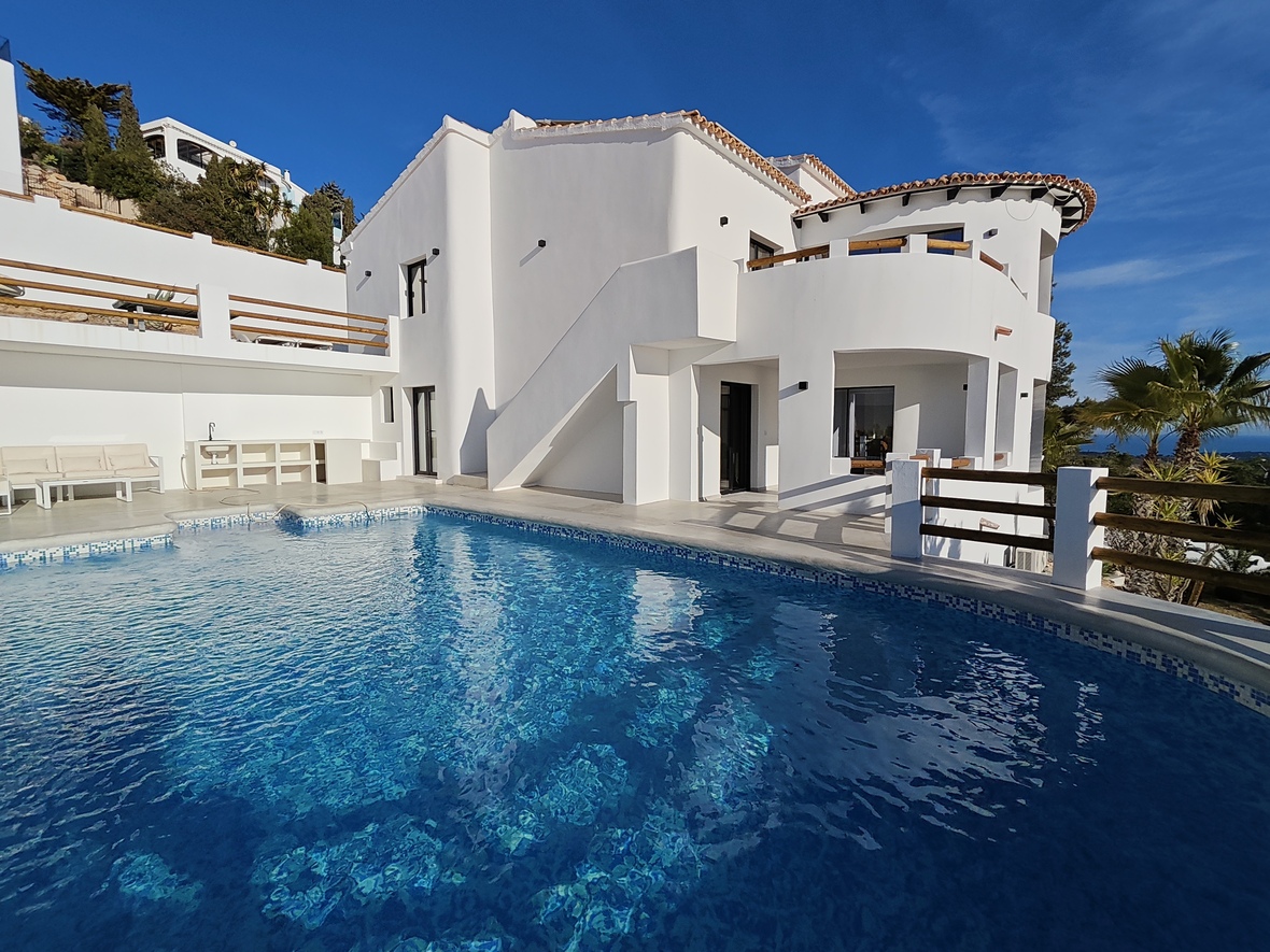 Vista exterior de una villa blanca con piscina curva de gresite azul, terrazas de varios niveles y barandillas de madera natural.