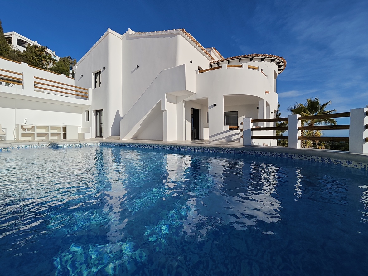 Vista exterior de una villa blanca con terrazas curvas, barandillas de madera y una piscina de gresite azul que refleja la arquitectura mediterránea.