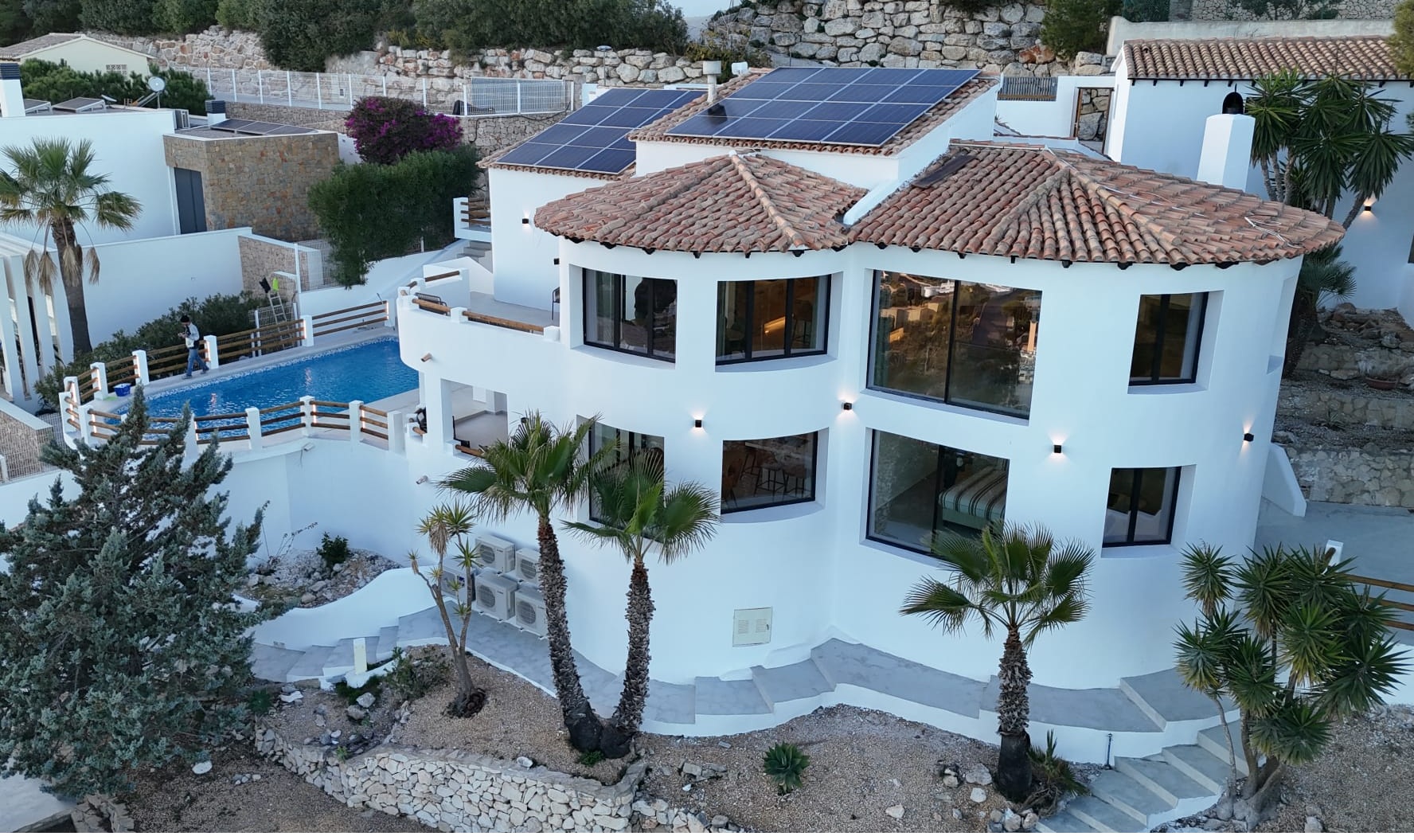 Vista exterior de una villa blanca de varios niveles con arquitectura curva, techos de teja con paneles solares y piscina privada rodeada de palmeras.