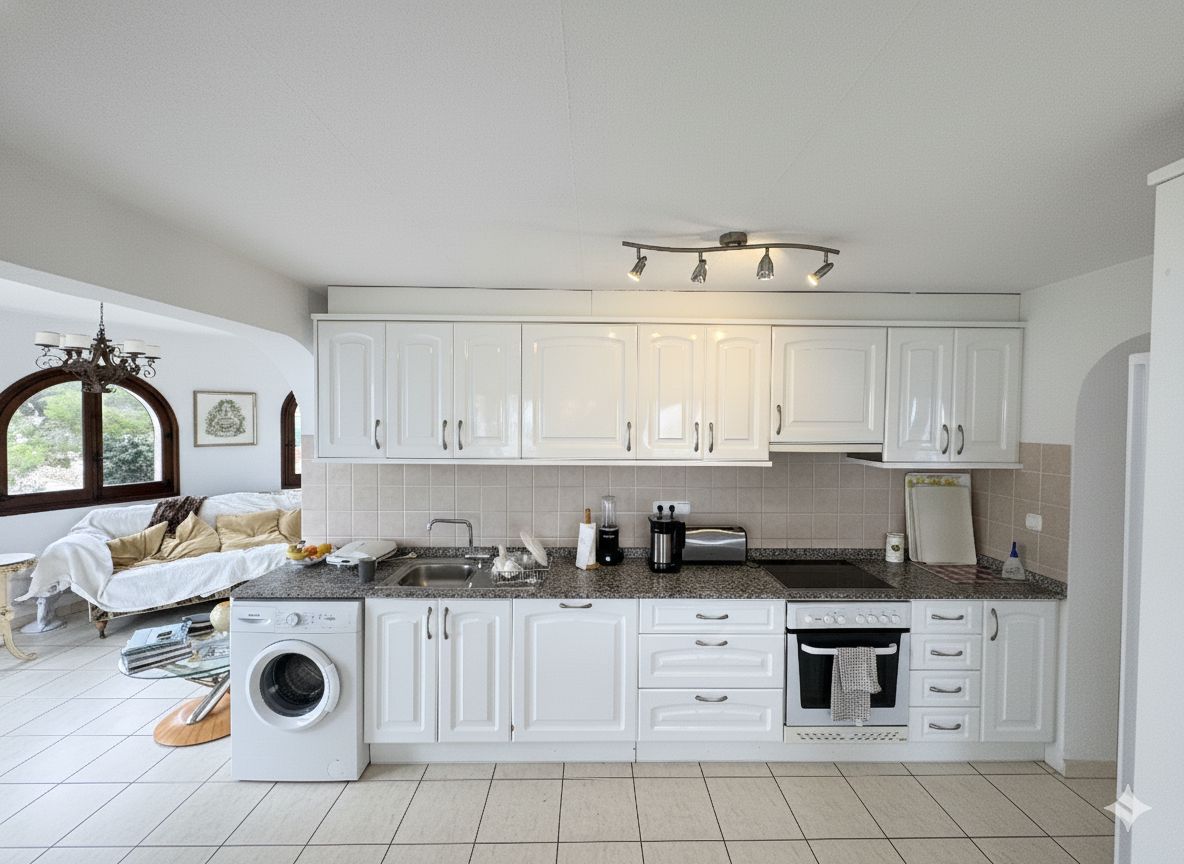 Cocina lineal con armarios blancos, encimera de granito y electrodomésticos integrados. Incluye lavadora, horno y un salpicadero de azulejos en tonos beige.