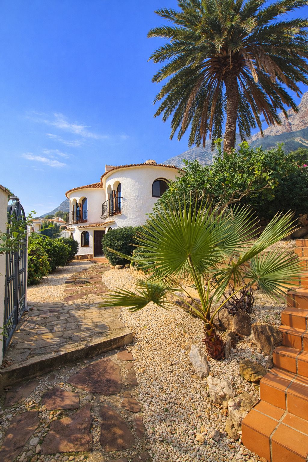Fachada de una villa mediterránea blanca con torres redondeadas, ventanas de arco y un camino de piedra rodeado de palmeras y vegetación autóctona frente a la montaña.