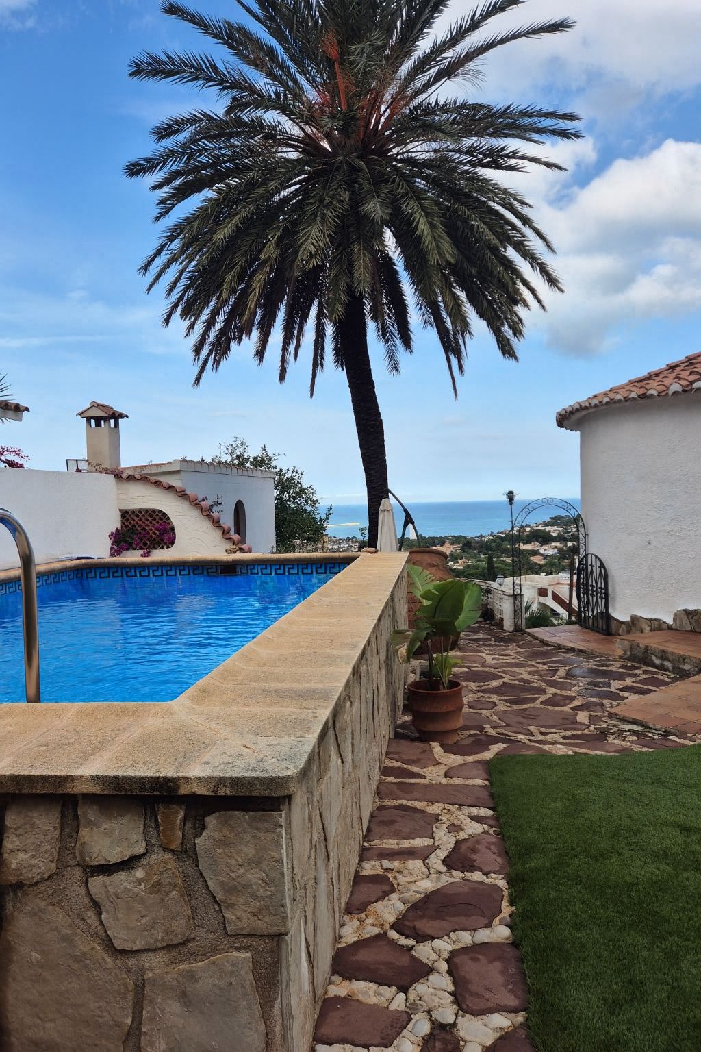 Piscina elevada con revestimiento de piedra natural, terraza de lajas, una gran palmera y vistas panorámicas hacia la costa mediterránea.