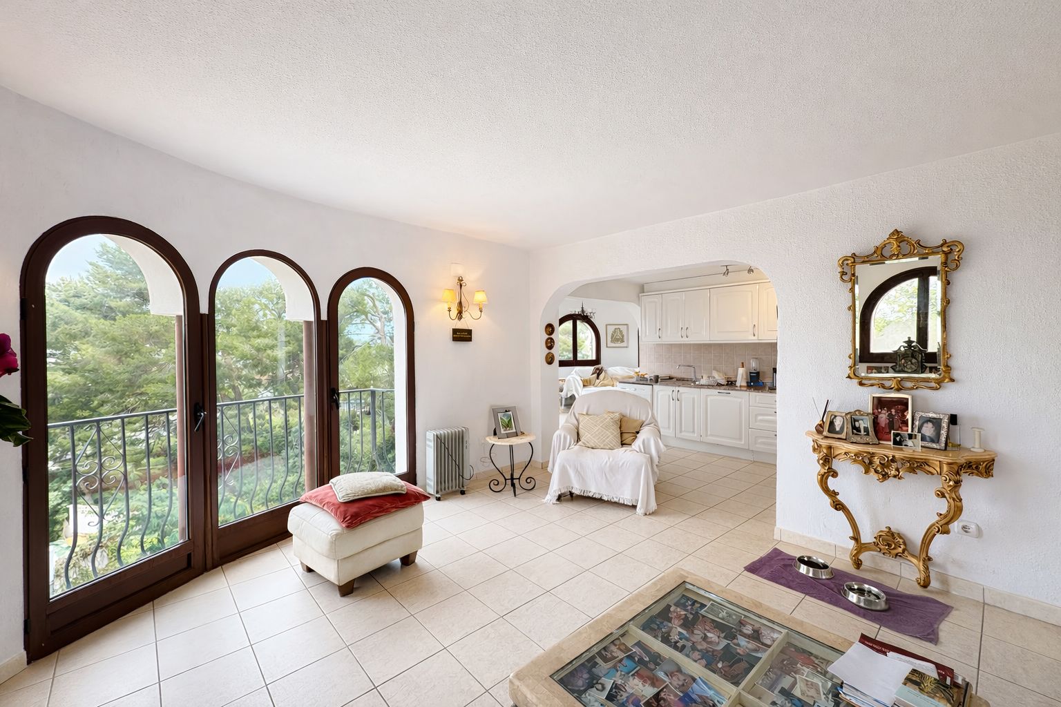 Salón luminoso con tres ventanales de arco, acceso a balcón con vistas arboladas, suelos cerámicos claros y mobiliario clásico con detalles dorados.