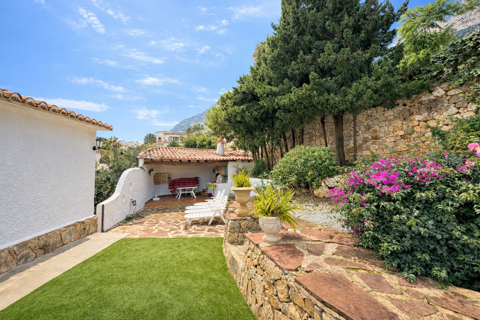 Terraza exterior con pavimento de piedra natural, césped artificial y porche cubierto con teja árabe. Incluye muros de mampostería y vegetación mediterránea.