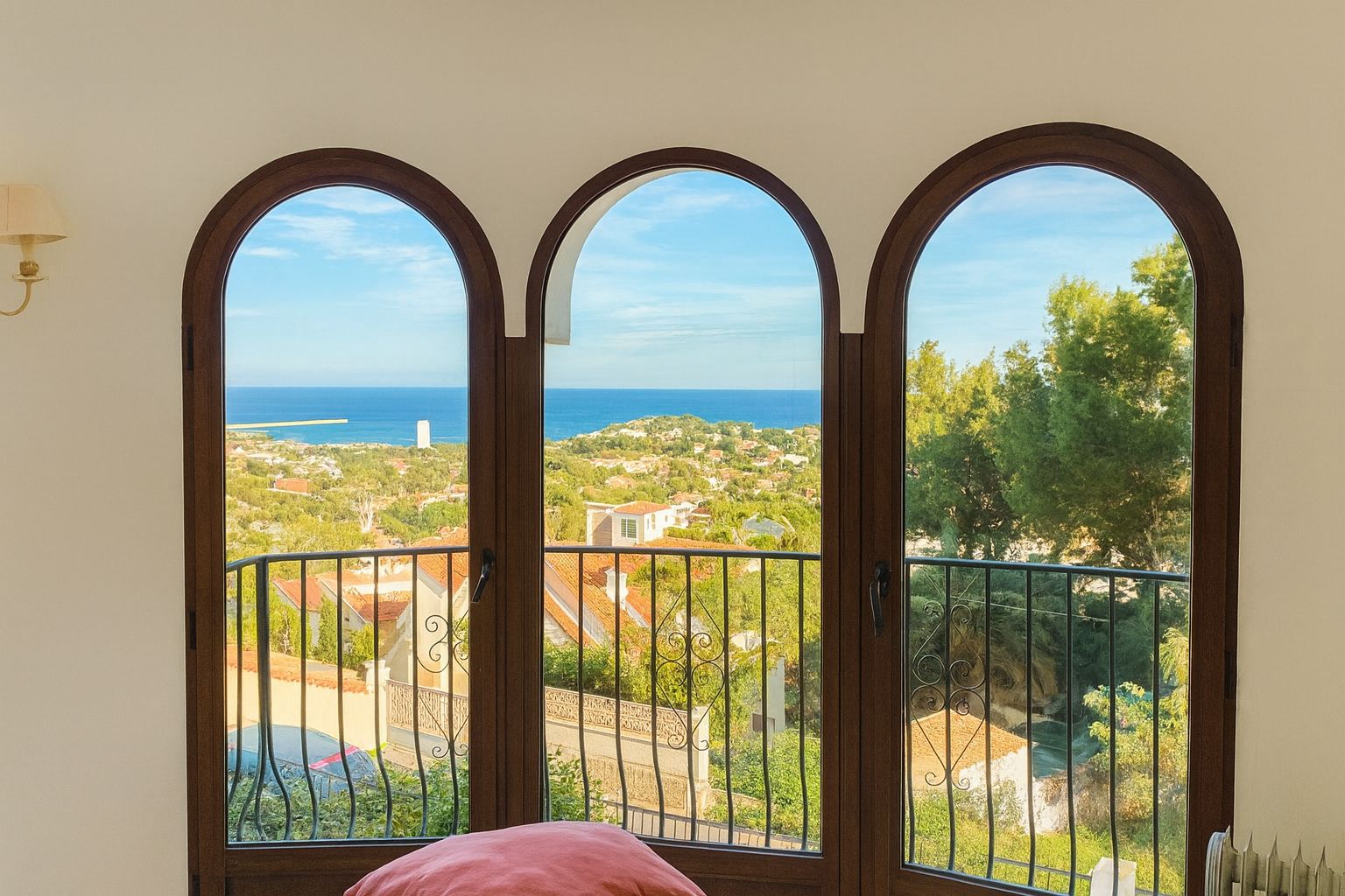 Tres ventanales de arco con marcos de madera oscura ofrecen vistas panorámicas al mar Mediterráneo y al paisaje costero, complementados por una barandilla de hierro forjado.