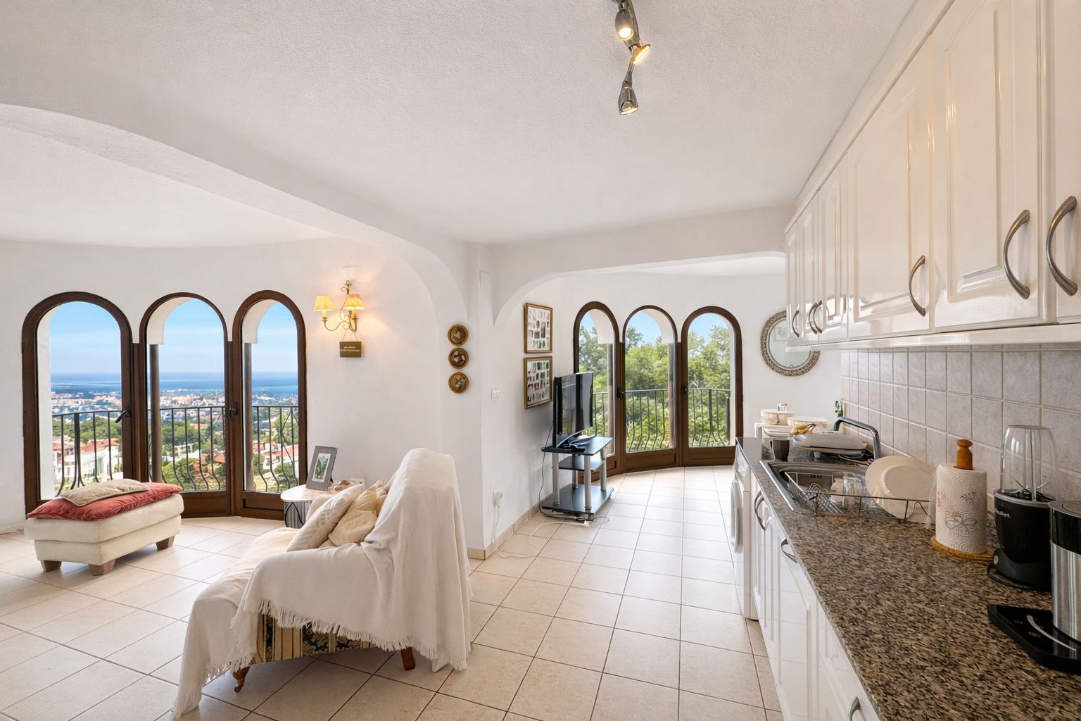 Zona de estar de planta abierta con cocina equipada, encimeras de granito y ventanales de medio punto con vistas panorámicas al mar y la costa.
