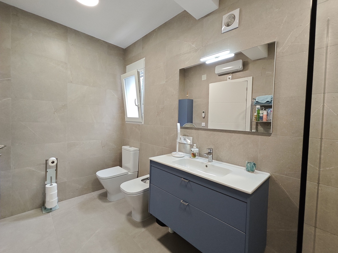 Baño moderno con revestimiento cerámico en tono arena, mueble suspendido azul con lavabo integrado, bidé e inodoro. Destaca el amplio espejo con iluminación LED superior.