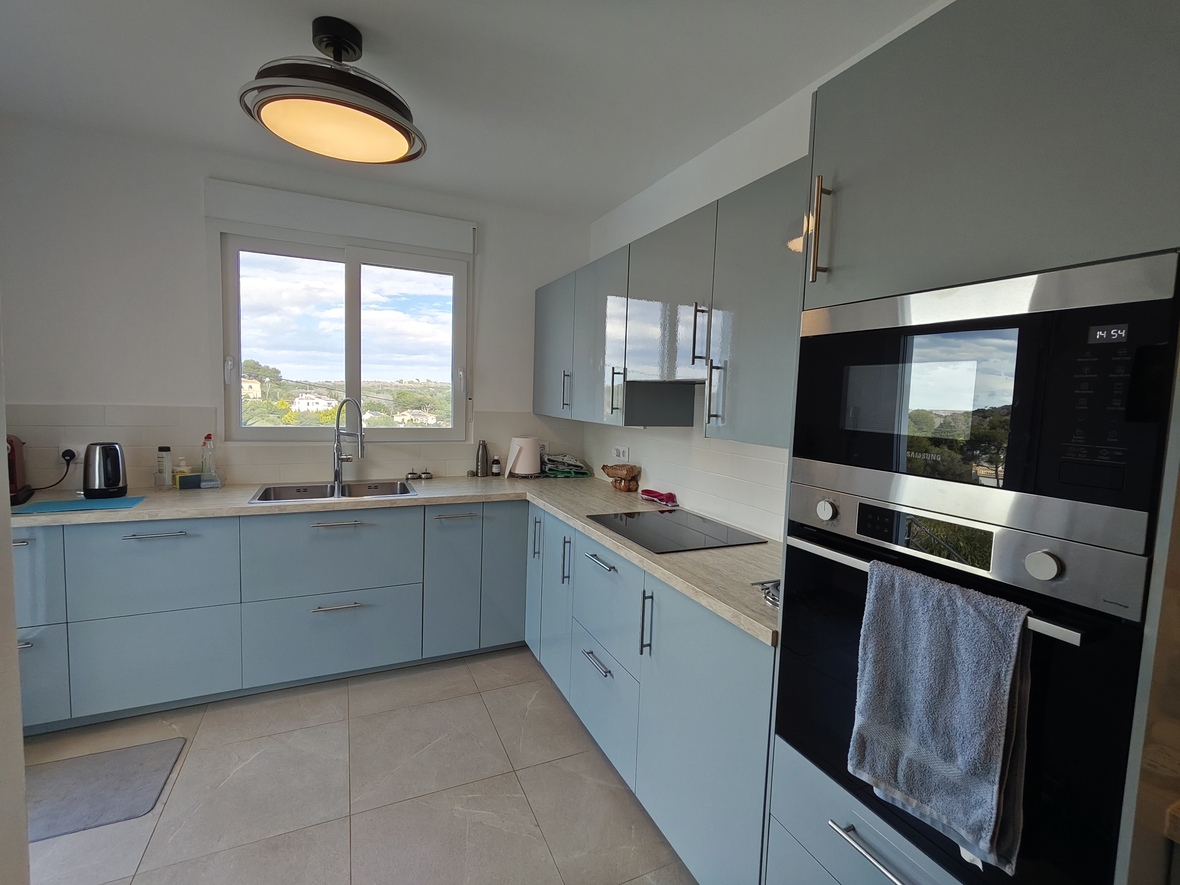 Cocina moderna con armarios en azul pálido, encimera de efecto madera y electrodomésticos Samsung integrados. El ventanal ofrece vistas despejadas al entorno natural.