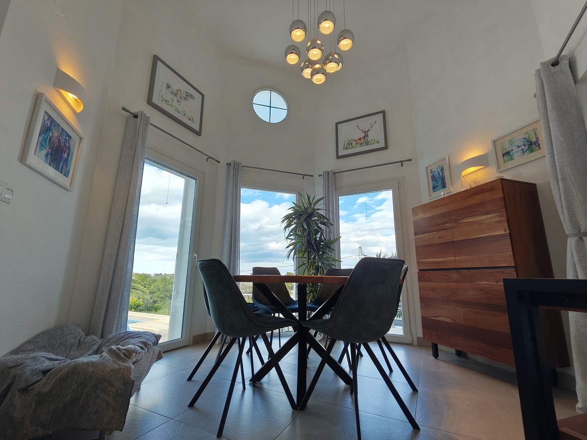 Comedor de techos altos con lámpara de esferas cromadas, óculo decorativo y aparador de madera maciza. Los ventanales ofrecen luz natural y vistas al entorno exterior.