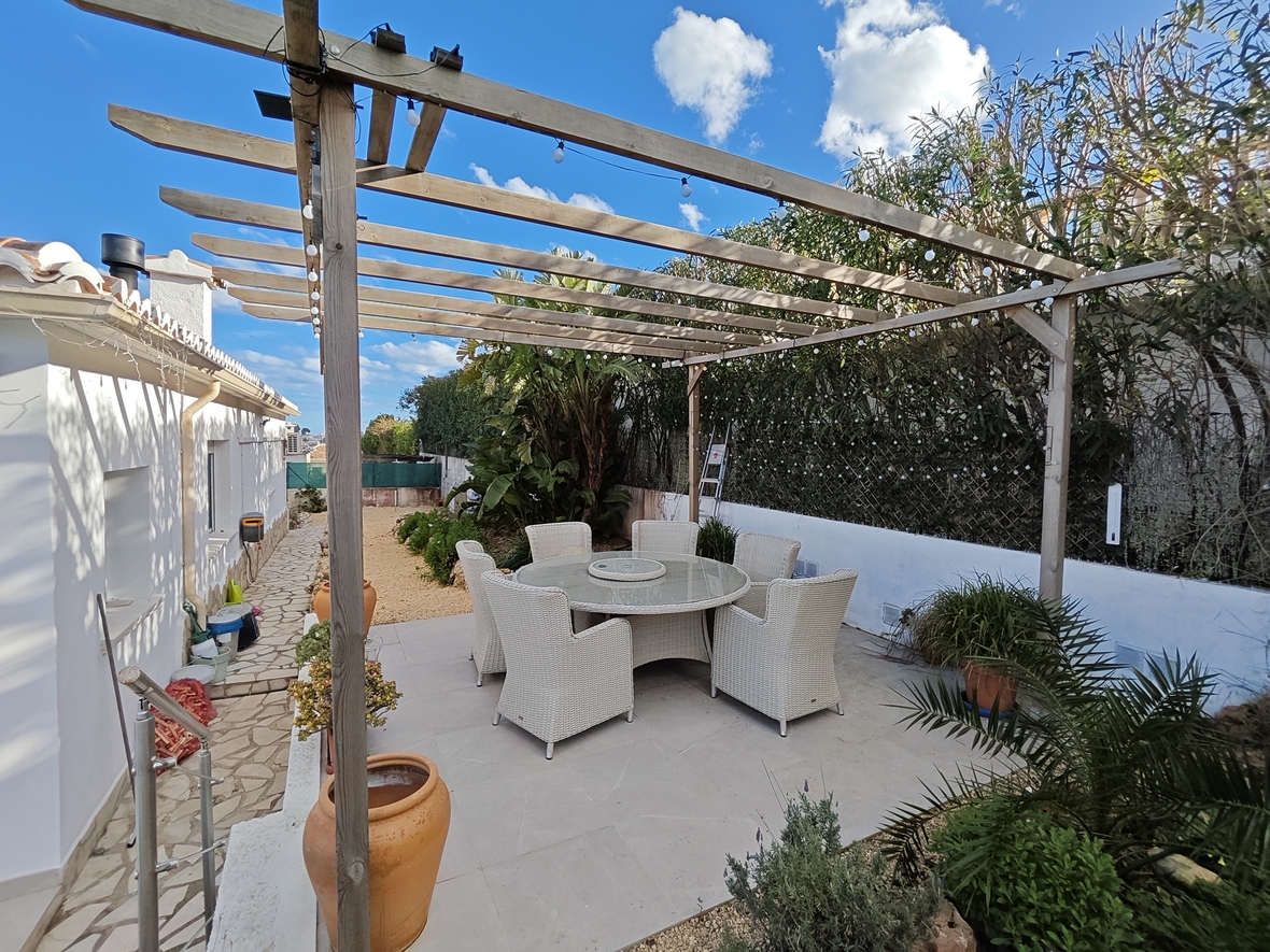 Comedor exterior bajo pérgola de madera con mesa circular de cristal y sillas de mimbre blanco, rodeado de vegetación mediterránea y macetas de terracota.