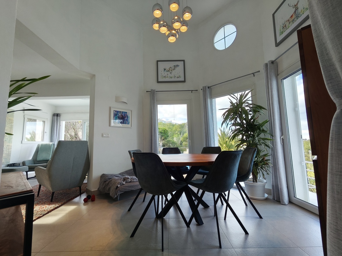 Comedor moderno con mesa redonda de madera, sillas tapizadas oscuras y techos altos con una ventana circular y una lámpara de araña cromada.