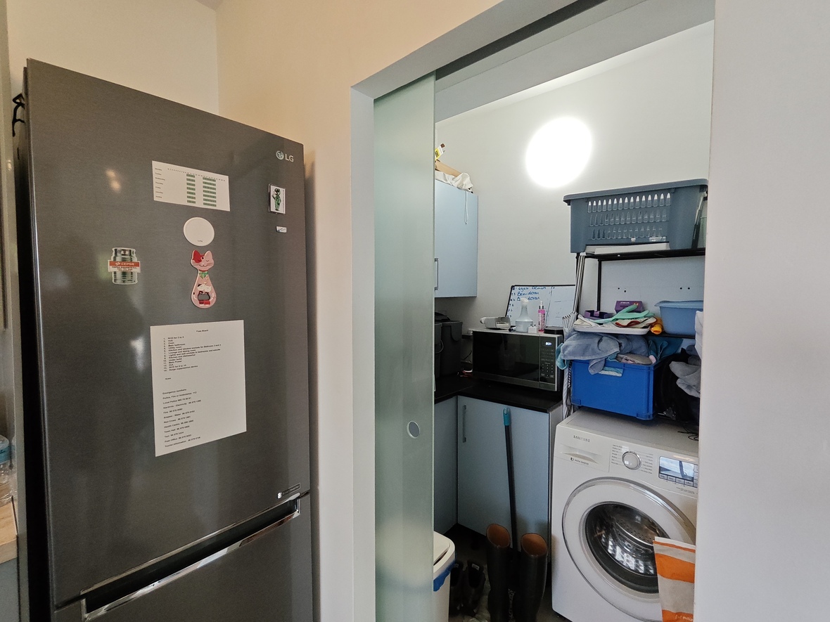 Cuarto de servicio con puerta corredera de cristal traslúcido, equipado con lavadora Samsung, microondas y armarios azul claro junto a un frigorífico LG.