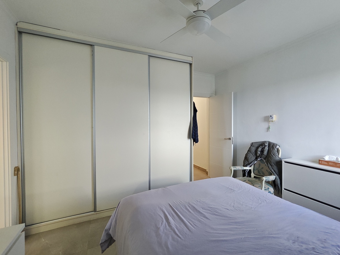 Dormitorio con amplio armario empotrado de tres puertas correderas blancas, ventilador de techo y mobiliario funcional en tonos claros.