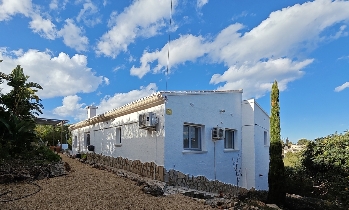 Fachada exterior blanca con zócalo de piedra natural, camino de grava y vegetación mediterránea bajo un cielo despejado.