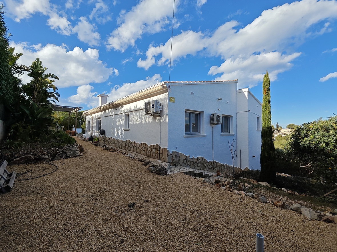 Fachada lateral de villa blanca con zócalo de piedra natural, camino de grava y unidades de aire acondicionado, rodeada de vegetación mediterránea y un ciprés.