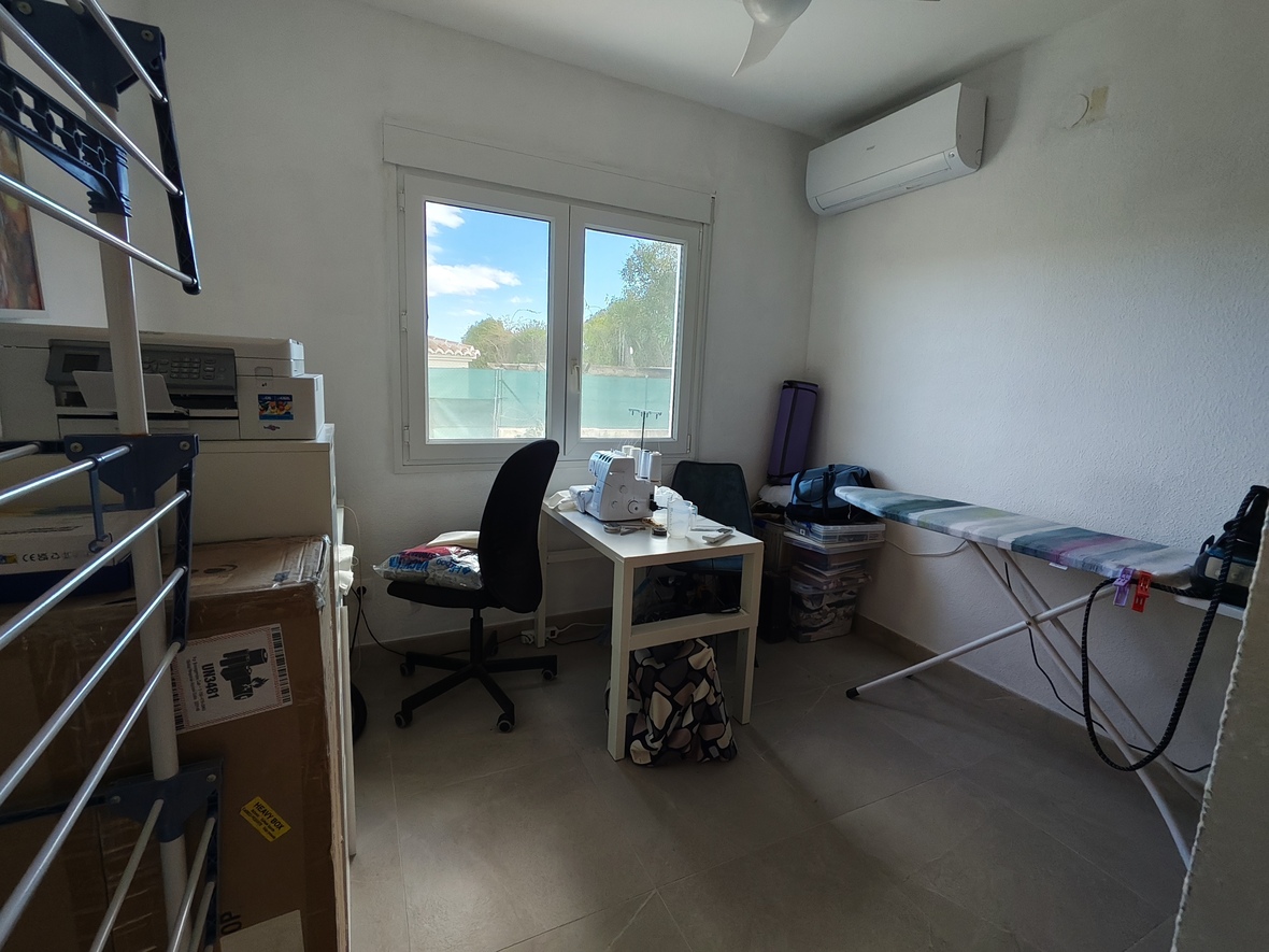 Habitación multiusos funcional con zona de costura, tabla de planchar y aire acondicionado. El espacio cuenta con suelos cerámicos y luz natural desde una ventana doble.