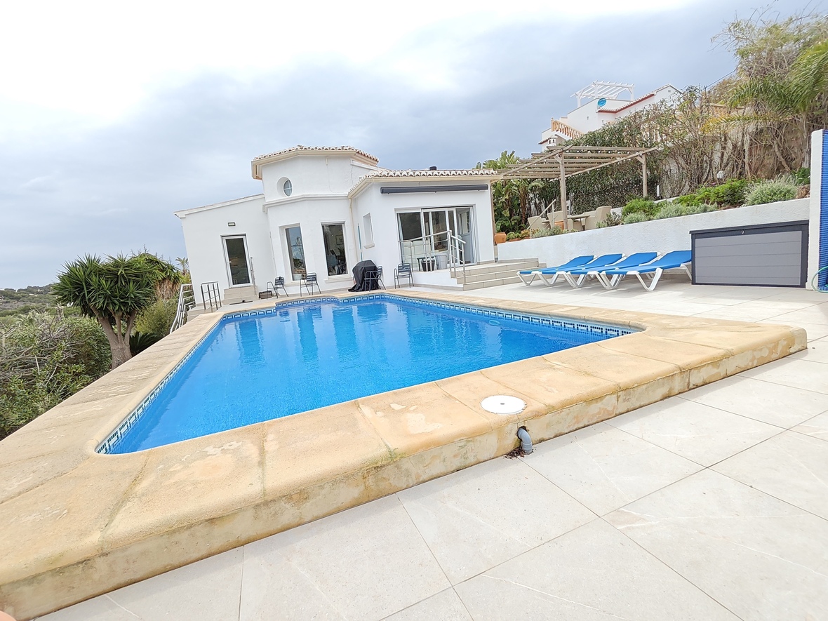 Piscina privada rectangular con revestimiento azul y borde de piedra, rodeada por una terraza de baldosas claras, tumbonas y una villa blanca de estilo mediterráneo.