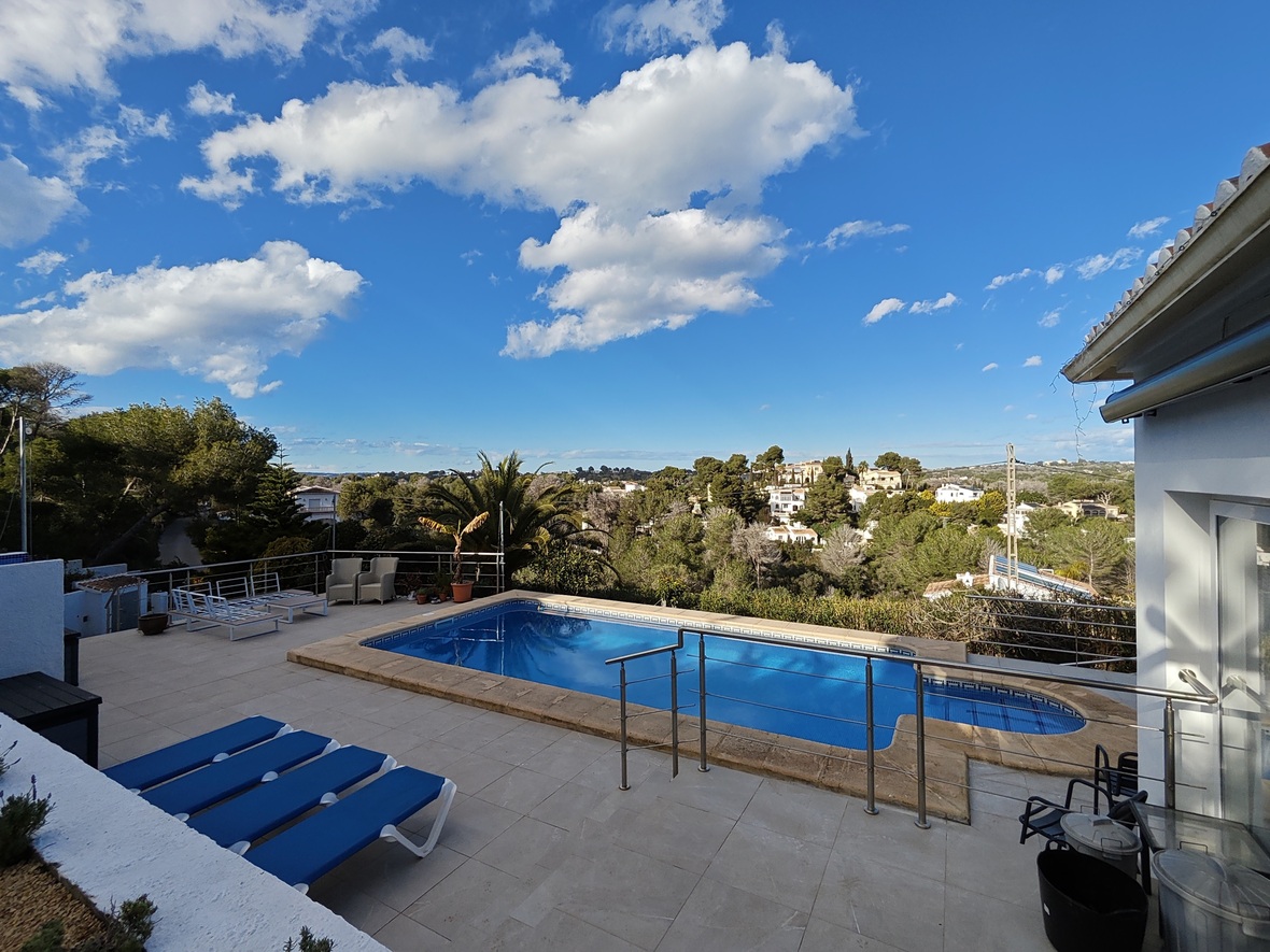Terraza elevada con piscina de gresite azul, borde de piedra y vistas despejadas hacia el valle residencial rodeado de pinos y palmeras.