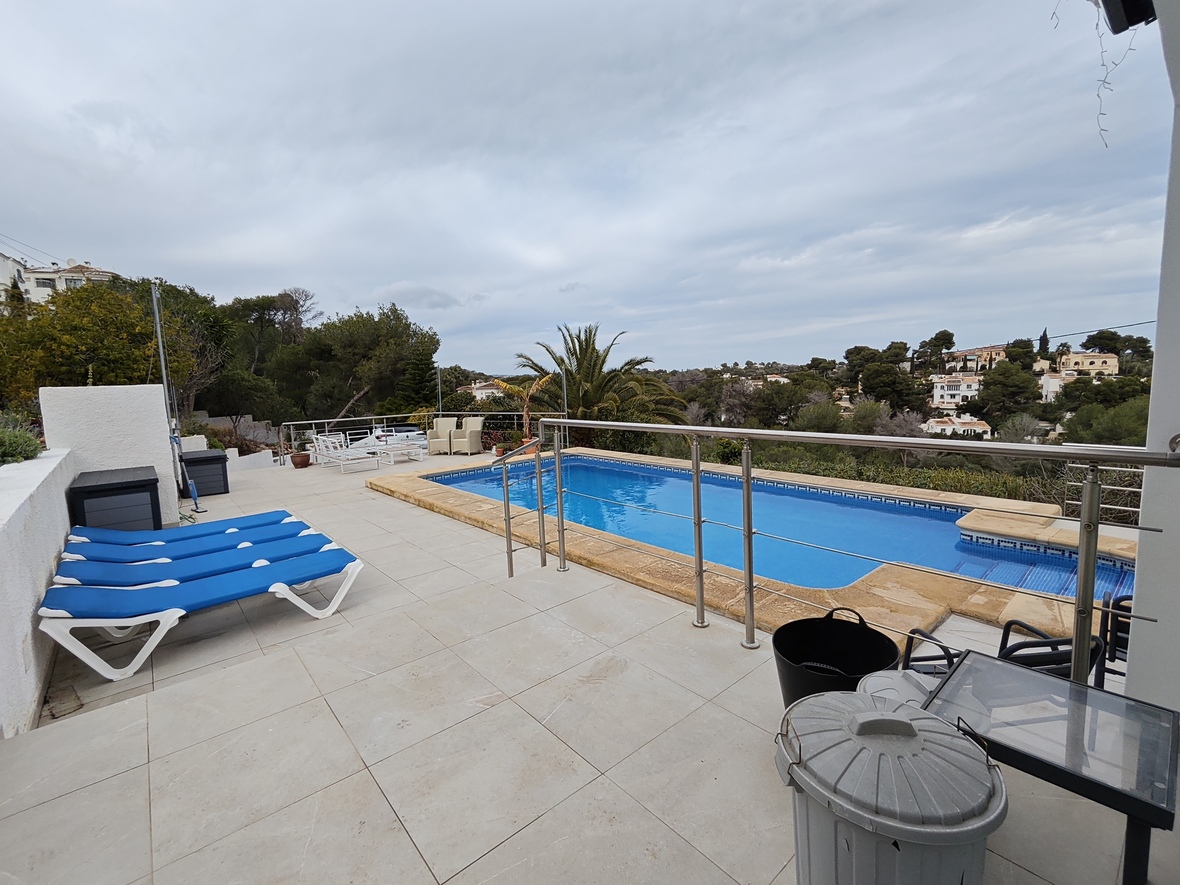 Terraza elevada con piscina rectangular, borde de piedra, tumbonas azules y barandillas de acero inoxidable con vistas a un entorno residencial arbolado.