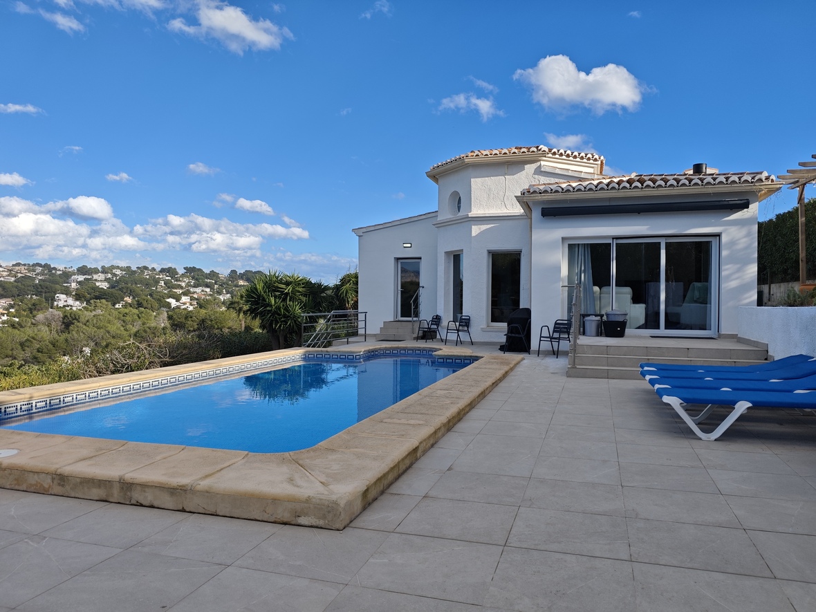 Terraza exterior con piscina rectangular, borde de piedra, tumbonas azules y vistas despejadas a las colinas y vegetación mediterránea.