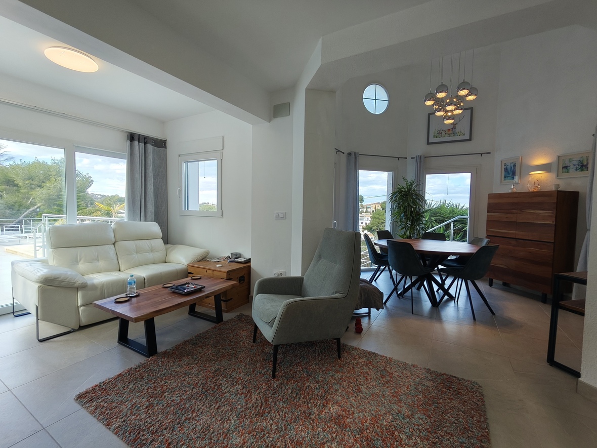 Zona de estar y comedor de concepto abierto con sofá de piel blanca, detalles en madera y lámpara de diseño. Amplios ventanales conectan con la terraza y ofrecen vistas despejadas.