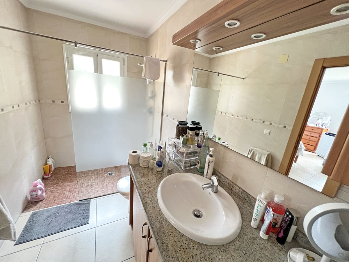Baño con encimera de granito, lavabo encastrado y espejo con iluminación integrada. Dispone de ducha a ras de suelo con mampara translúcida y suelo de mosaico.