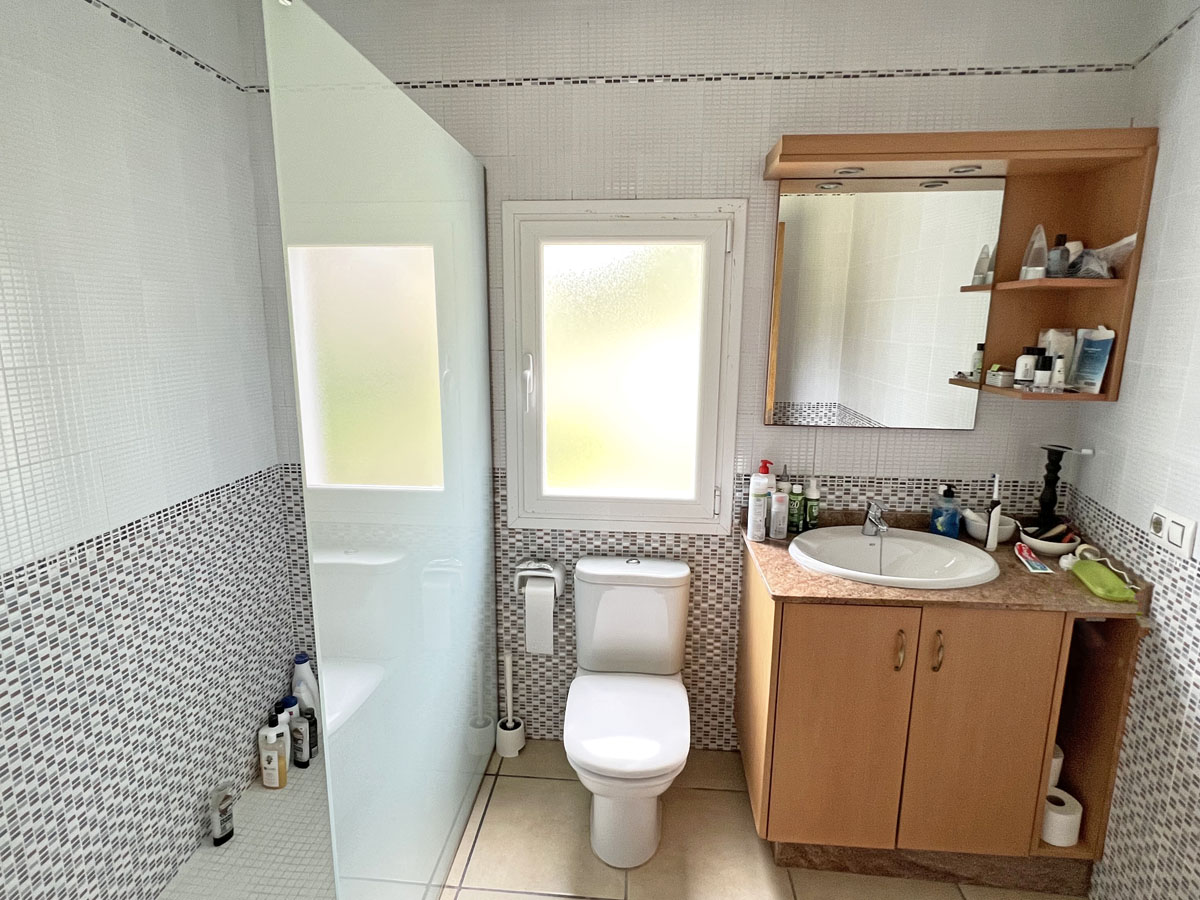 Baño funcional con ducha de obra y mampara de cristal translúcido, mueble de lavabo en madera con encimera de mármol y paredes revestidas en mosaico cerámico.