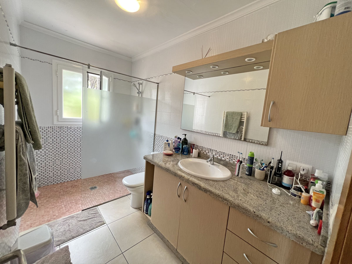 Cuarto de baño con encimera de granito, mueble de madera clara y espejo con focos integrados. Dispone de una ducha de obra con mampara de cristal translúcido y azulejos decorativos.