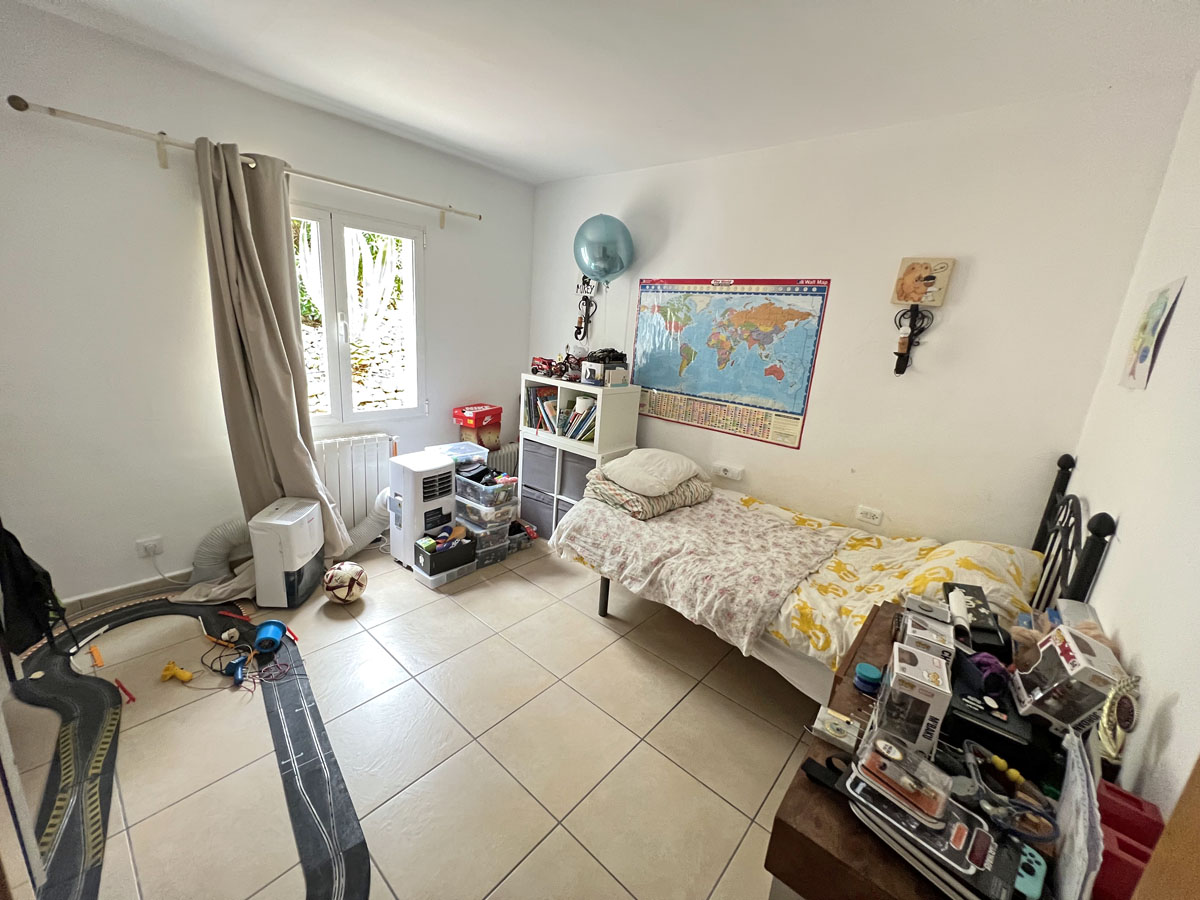 Dormitorio luminoso con suelos de gres, cama individual y ventana al exterior. Incluye estanterías blancas, radiador y paredes en tonos neutros con luz natural.