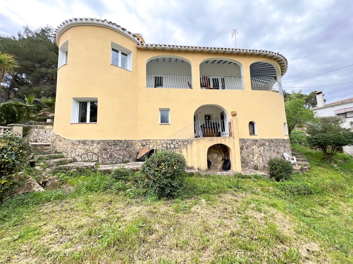 Fachada curva de estilo mediterráneo con zócalo de piedra natural, terrazas cubiertas en la planta superior y jardín en pendiente con vegetación autóctona.